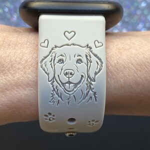 K&ouml;nnte beinhalten: Hellgraues Armband mit detaillierter Gravur eines Hundegesichts, umgeben von Herzen und Pfotenabdr&uuml;cken. Das Armband ist an einer schwarzen Uhr befestigt.