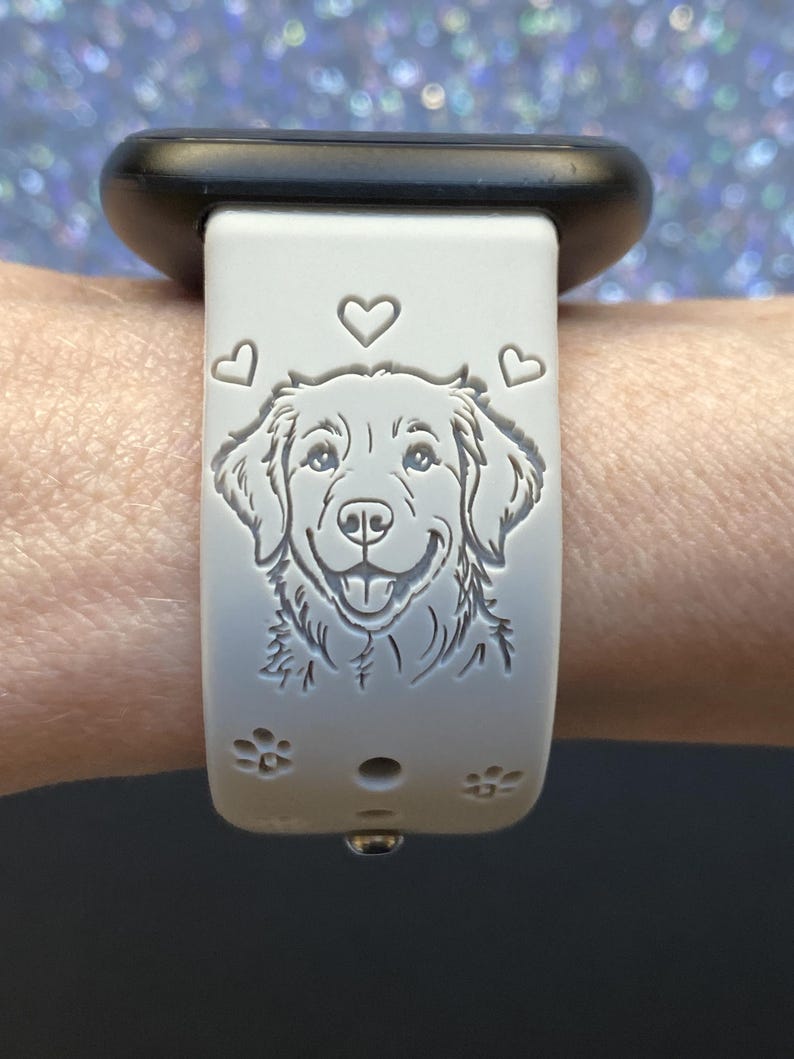 K&ouml;nnte beinhalten: Hellgraues Armband mit detaillierter Gravur eines Hundegesichts, umgeben von Herzen und Pfotenabdr&uuml;cken. Das Armband ist an einer schwarzen Uhr befestigt.