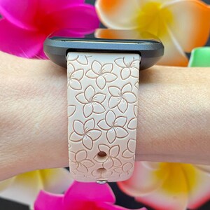 May include: A black smartwatch with a pink silicone band featuring a floral pattern. The band has a textured surface with raised flower designs.