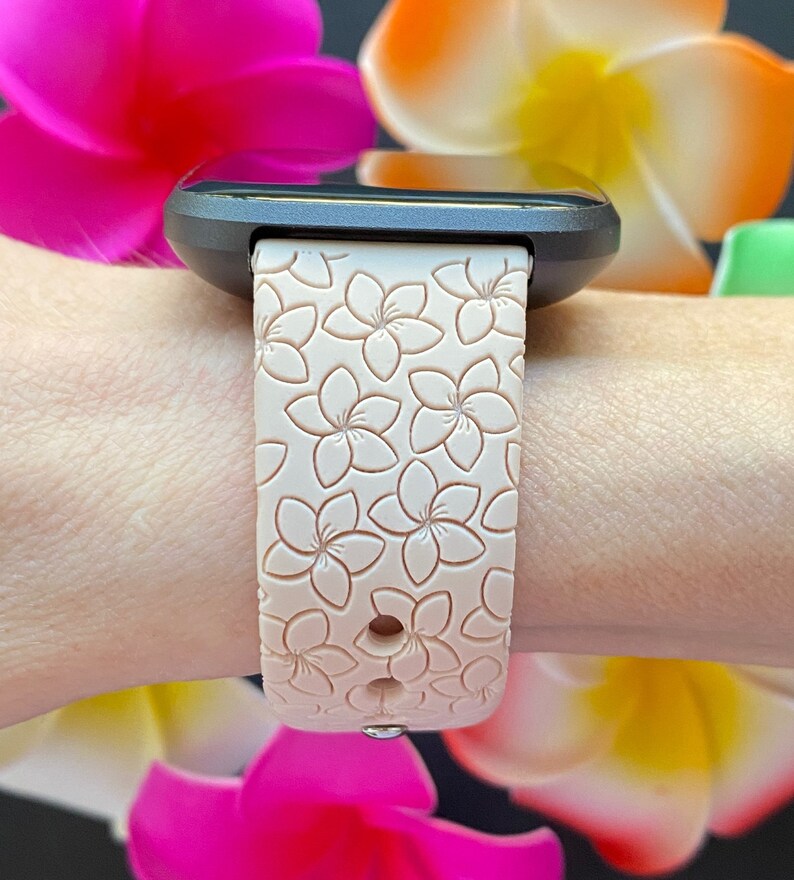 May include: A black smartwatch with a pink silicone band featuring a floral pattern. The band has a textured surface with raised flower designs.