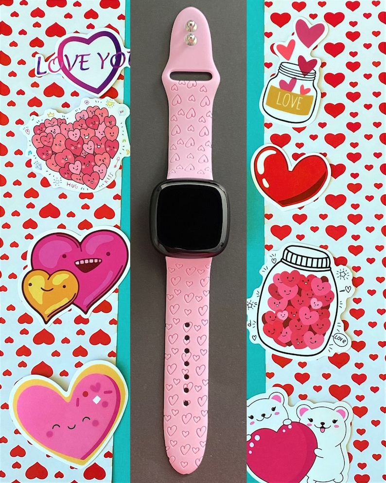 May include: A pink smartwatch with a black face and a pink band featuring a heart pattern. The band has a heart pattern and the watch face is black. The watch is surrounded by stickers with hearts and the words "Love You" and "Love".