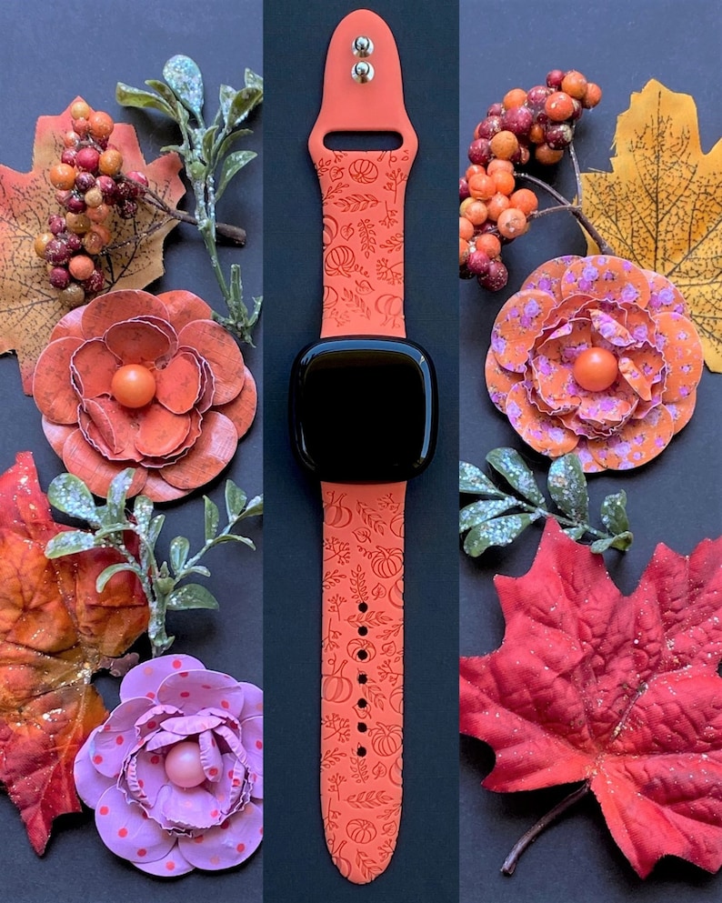 May include: A bright orange silicone Apple Watch band with a fall-themed design featuring pumpkins, leaves, and vines. The band is shown on a black background with fall-themed decorations.