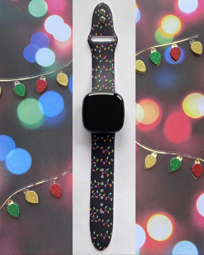 May include: Black smartwatch with a festive Christmas lights pattern on the band. The band features a repeating design of colorful lights on a dark background. The watch is displayed against a backdrop of blurred lights and decorative ornaments.