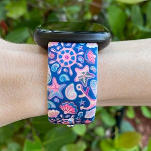 K&ouml;nnte beinhalten: Ein blaues und pinkfarbenes Armband mit nautischem Muster. Das Armband zeigt Muscheln, Anker und Seesterne.