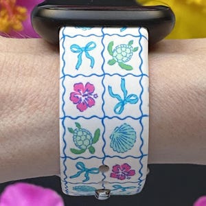 Op de afbeelding: Witte horlogeband met een herhaald patroon van blauwe strikken, roze hibiscusbloemen, groene schildpadden en een blauwe zeeschelp. Bevestigd aan een zwarte smartwatch.