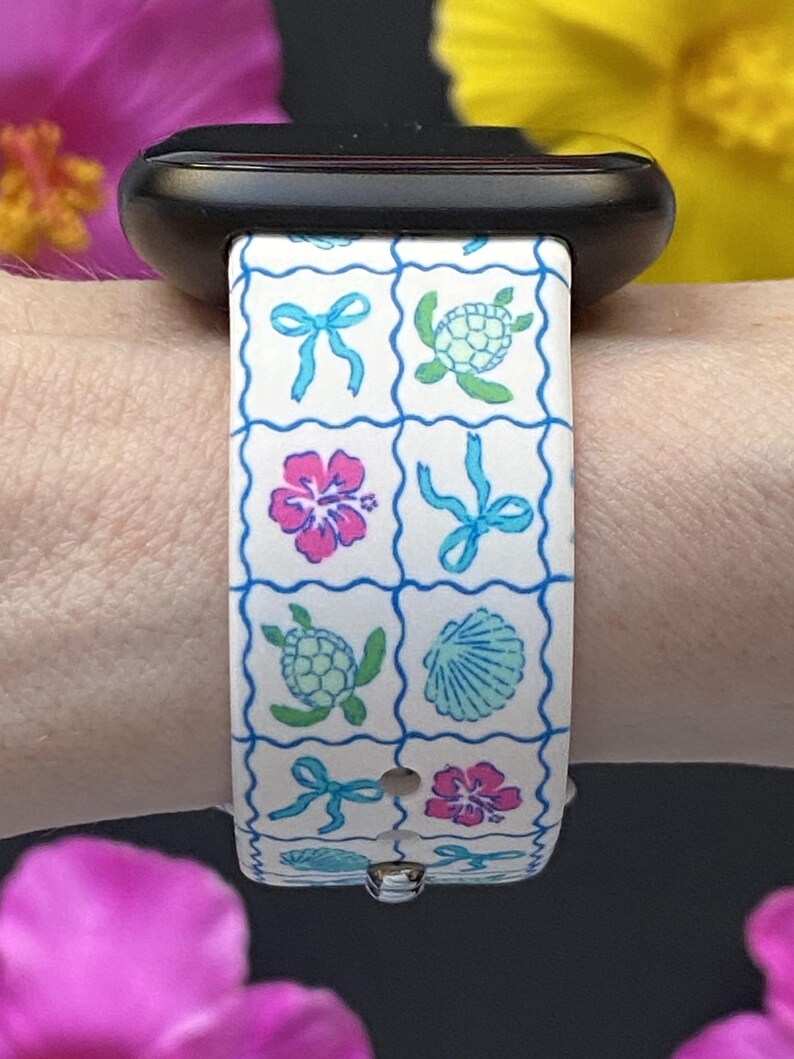 Op de afbeelding: Witte horlogeband met een herhaald patroon van blauwe strikken, roze hibiscusbloemen, groene schildpadden en een blauwe zeeschelp. Bevestigd aan een zwarte smartwatch.