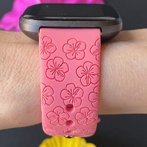 May include: A pink silicone watch band with a floral pattern. The band is designed for a smartwatch and has a black metal clasp.