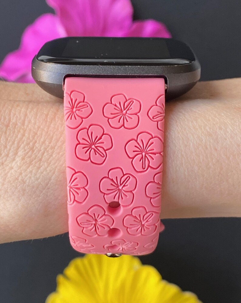 May include: A pink silicone watch band with a floral pattern. The band is designed for a smartwatch and has a black metal clasp.