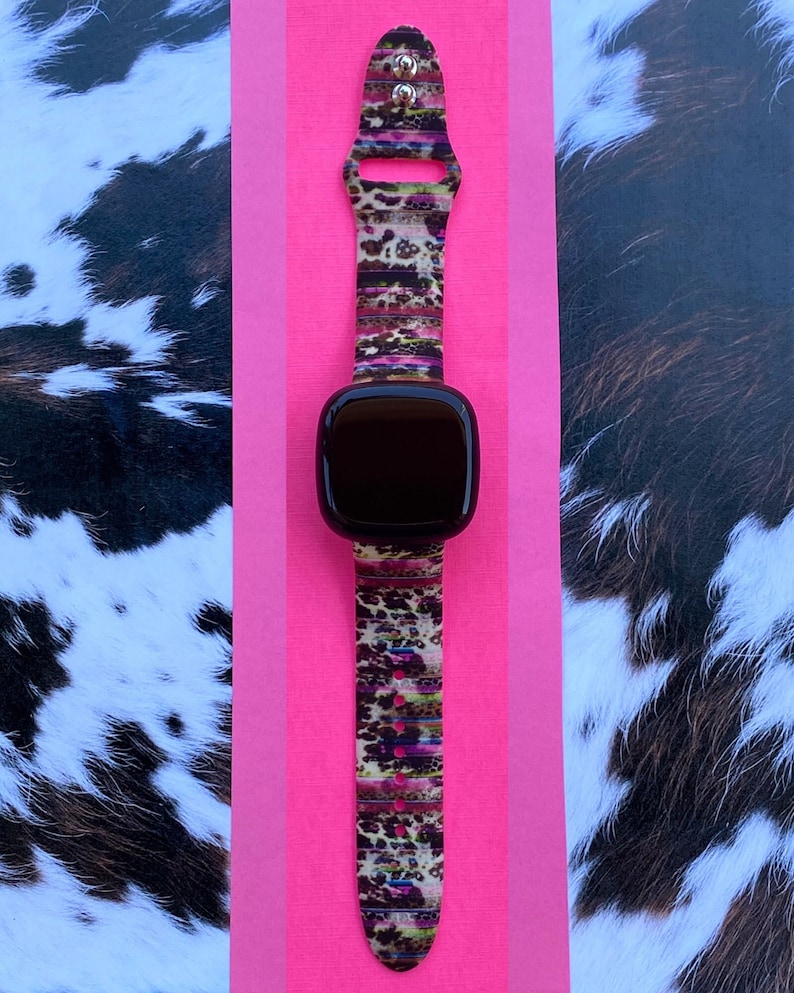 May include: A black smartwatch with a colorful leopard print band. The band has a pink, brown, and green pattern. The watch is on a pink background.
