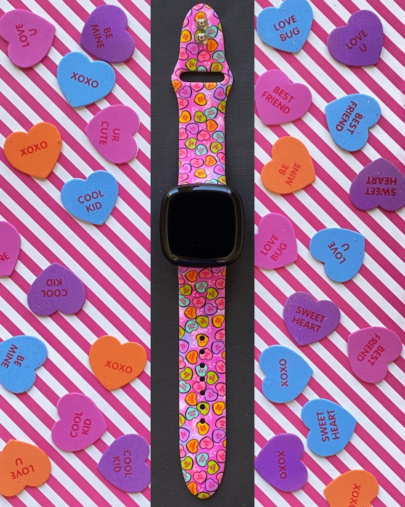 May include: A pink and white Apple Watch band with a heart-shaped pattern. The band is surrounded by various Valentine's Day themed candy hearts with messages like "Love Bug", "Best Friend", and "Sweet Heart".