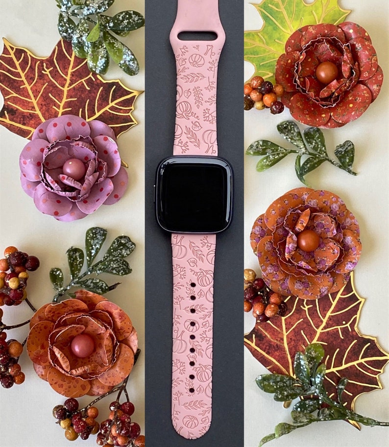K&ouml;nnte beinhalten: Ein rosafarbenes Apple Watch Armband mit einem Herbst-Design mit K&uuml;rbissen und Bl&auml;ttern. Das Armband wird vor einem Hintergrund aus herbstlichen Dekorationen gezeigt.