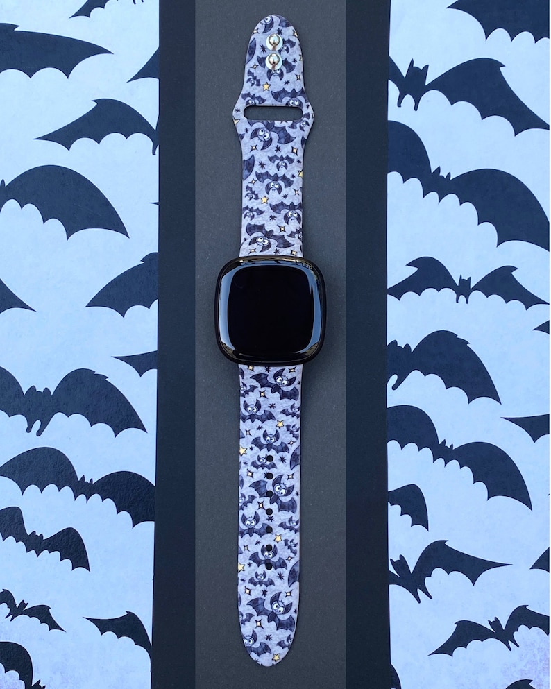 Puede incluir: Un reloj inteligente negro con una banda de color morado y negro con estampado de murci&eacute;lagos. La banda tiene peque&ntilde;as estrellas doradas.