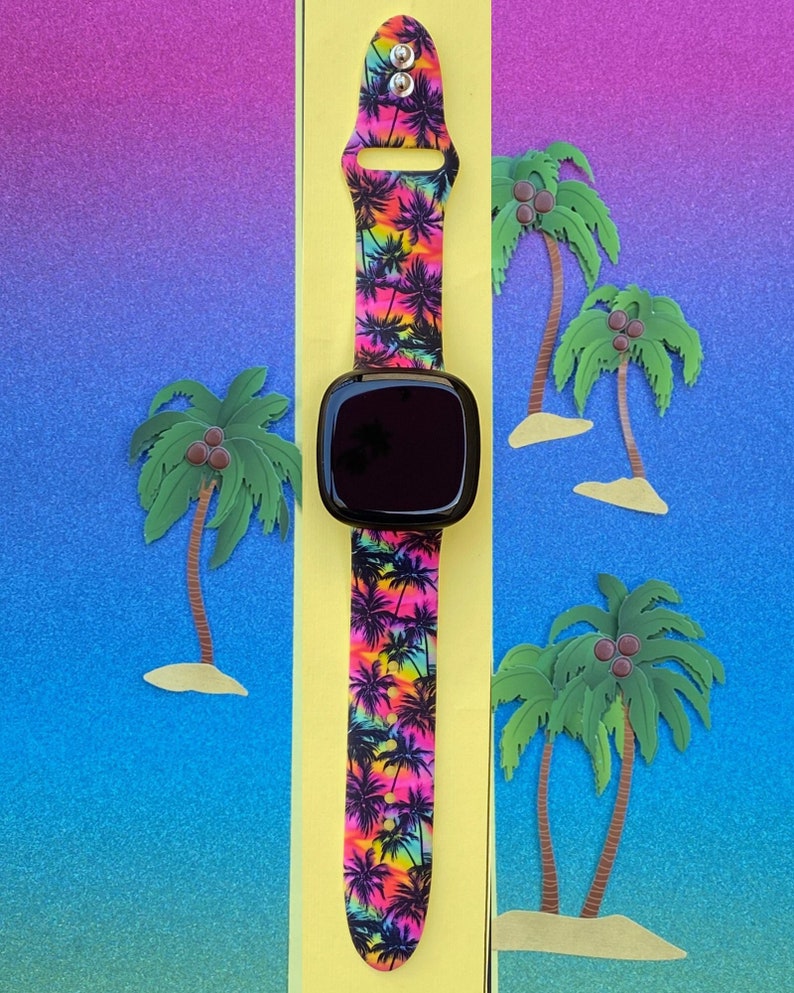 May include: A black smartwatch with a vibrant band featuring a tropical palm tree pattern in shades of pink, yellow, and black. The watch is displayed on a yellow background with paper cut-out palm trees and a gradient blue and purple backdrop.