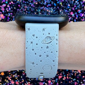 May include: A gray silicone watch band with a white star and planet design. The band is on a person's wrist.