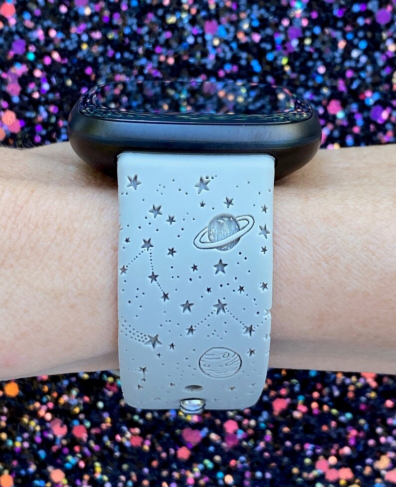 May include: A gray silicone watch band with a white star and planet design. The band is on a person's wrist.