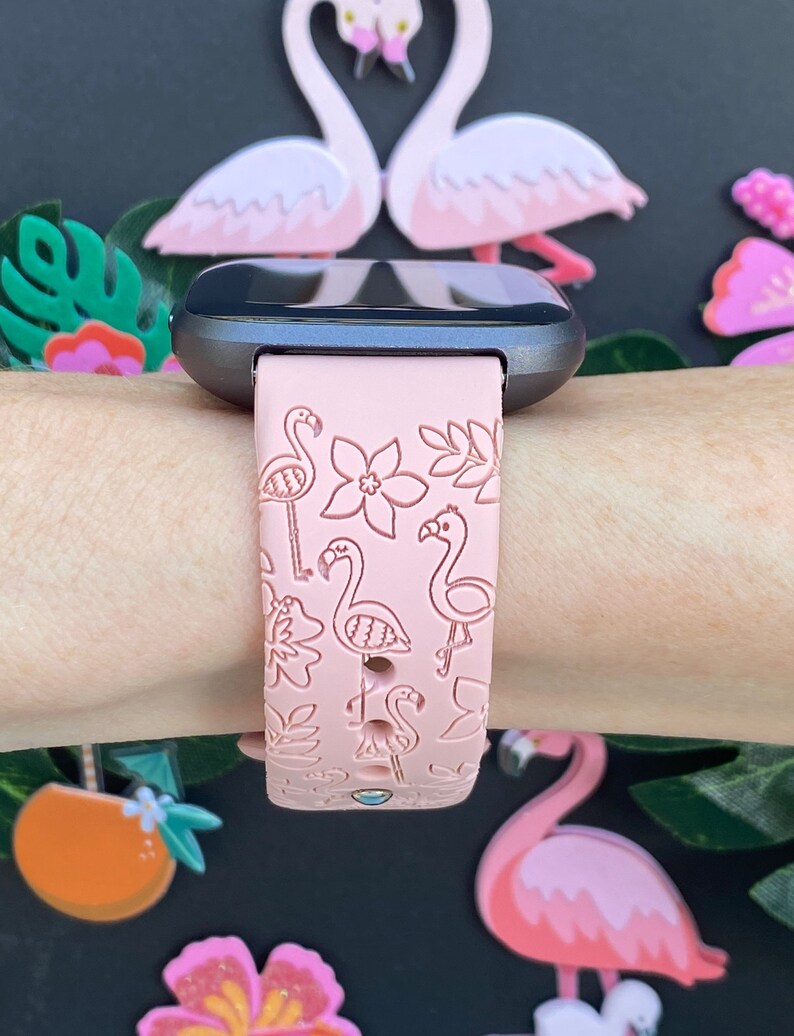 May include: A pink silicone watch band with a flamingo and floral pattern. The band is designed for a smartwatch and has a black watch face.