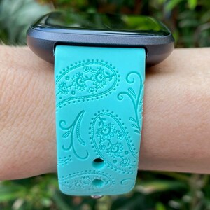May include: A turquoise blue smartwatch band with a paisley pattern. The band is made of silicone and has a black metal buckle.