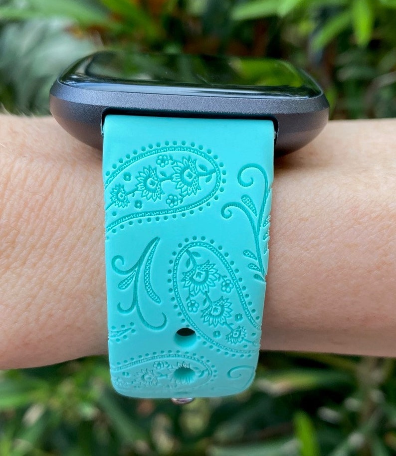 May include: A turquoise blue smartwatch band with a paisley pattern. The band is made of silicone and has a black metal buckle.