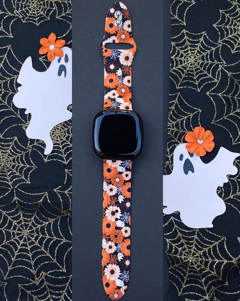 May include: A black smartwatch with a bright orange, white and black patterned band featuring ghosts and flowers. The band is made of silicone and has a buckle closure.