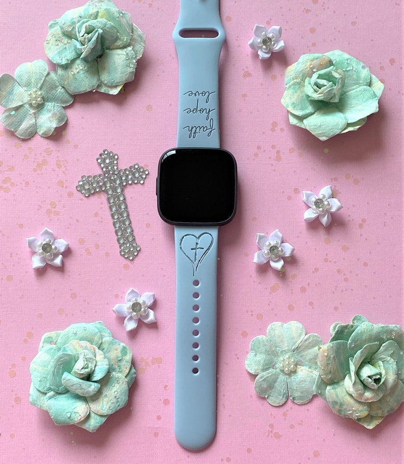 May include: A light blue watch strap with the words "Faith, Hope, Love" engraved on it. The strap has a small heart with a cross inside on the bottom. The watch strap is surrounded by small white flowers and green paper flowers.
