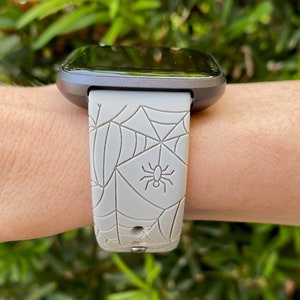 May include: A gray smartwatch with a spiderweb and spider design on the band.