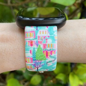 K&ouml;nnte beinhalten: Schwarze Smartwatch mit einem Armband mit einem Winterdorfmuster. Das Armband zeigt H&auml;user mit rosa, gelben und blauen Akzenten auf t&uuml;rkisfarbenem Hintergrund. Ein gr&uuml;ner Baum und ein violetter Baum sind ebenfalls zu sehen.