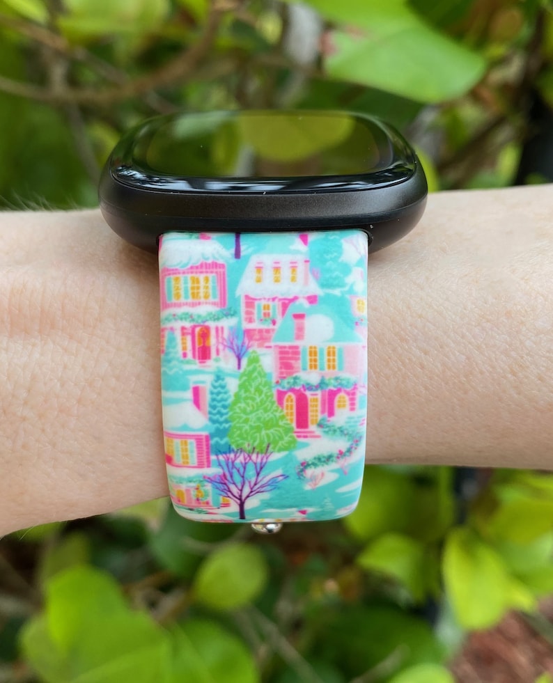K&ouml;nnte beinhalten: Schwarze Smartwatch mit einem Armband mit einem Winterdorfmuster. Das Armband zeigt H&auml;user mit rosa, gelben und blauen Akzenten auf t&uuml;rkisfarbenem Hintergrund. Ein gr&uuml;ner Baum und ein violetter Baum sind ebenfalls zu sehen.