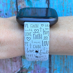 Puede incluir: Banda de reloj blanca con una esfera negra. La banda tiene un patr&oacute;n repetido de las palabras "fe", "amor" y "esperanza" con corazones y cruces.