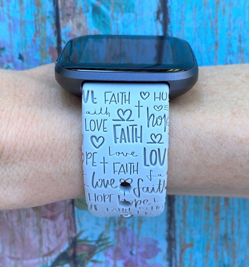 Puede incluir: Banda de reloj blanca con una esfera negra. La banda tiene un patr&oacute;n repetido de las palabras "fe", "amor" y "esperanza" con corazones y cruces.