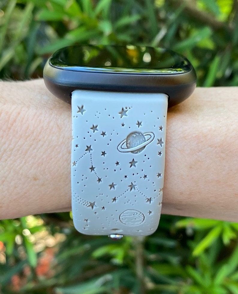 May include: A gray smartwatch band with a white background and a design of stars and planets. The band is made of silicone and has a smooth finish.