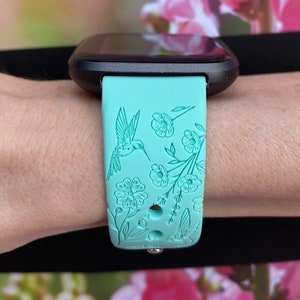 May include: A turquoise blue silicone watch band with a floral and hummingbird design. The band is designed for a smartwatch.