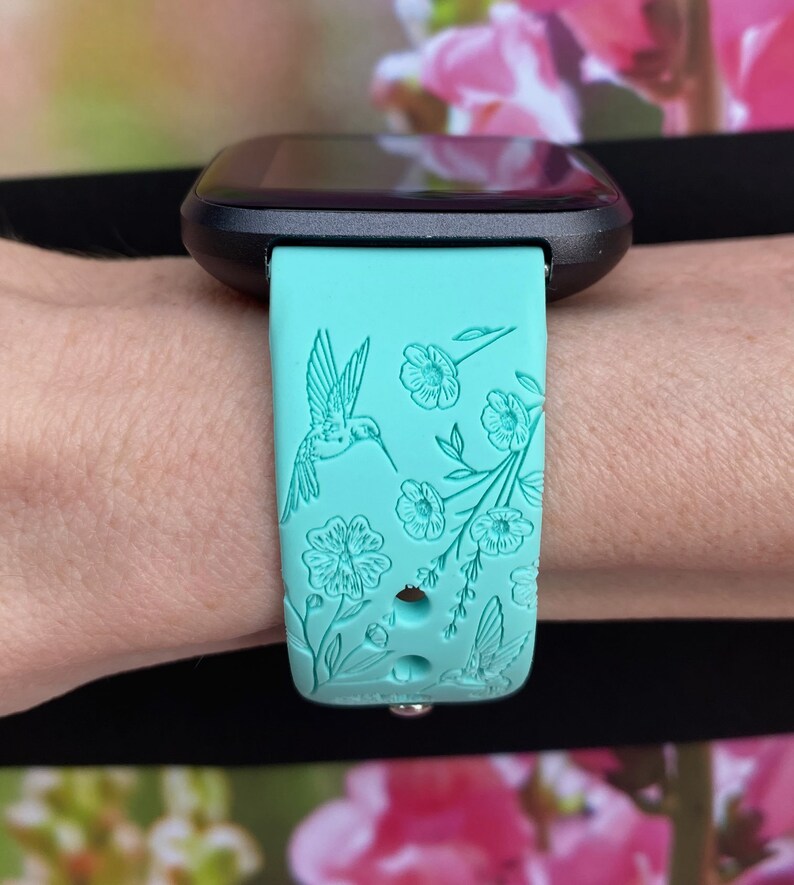 May include: A turquoise blue silicone watch band with a floral and hummingbird design. The band is designed for a smartwatch.