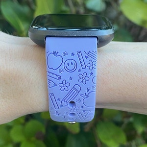 May include: A purple smartwatch with a black face and a purple band featuring a school-themed design with apples, pencils, rulers, and stars.