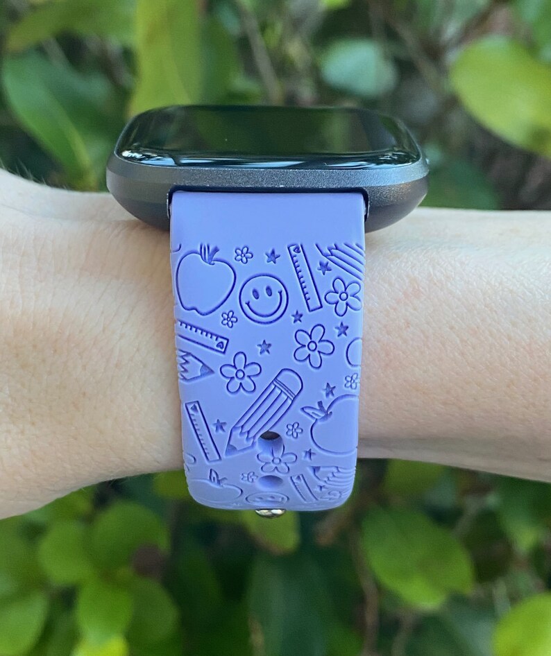 May include: A purple smartwatch with a black face and a purple band featuring a school-themed design with apples, pencils, rulers, and stars.