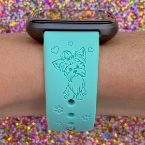 May include: A turquoise silicone watch band with a small, light blue Yorkshire Terrier dog design and three small hearts. The band has paw print accents and holes for adjusting the fit.