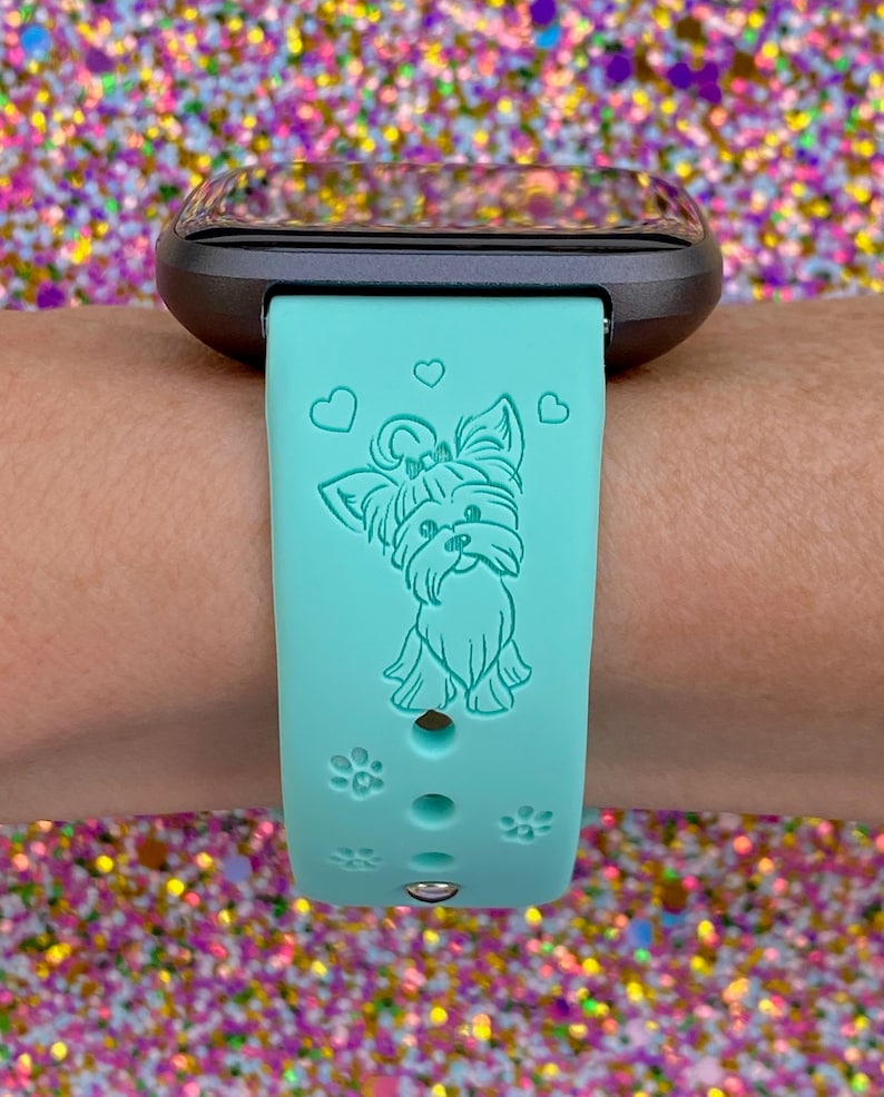 May include: A turquoise silicone watch band with a small, light blue Yorkshire Terrier dog design and three small hearts. The band has paw print accents and holes for adjusting the fit.