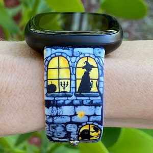 May include: A black smartwatch with a colorful Halloween-themed band. The band features a brick wall with a window and a witch inside. The window is lit with a yellow glow.