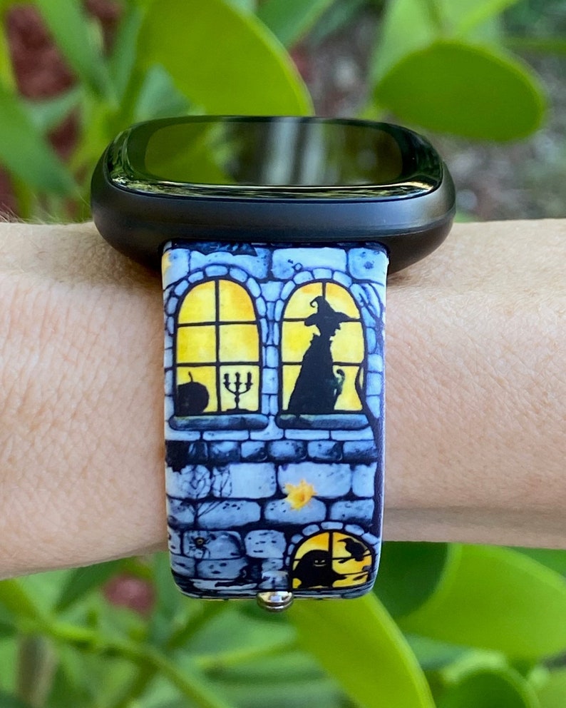 May include: A black smartwatch with a colorful Halloween-themed band. The band features a brick wall with a window and a witch inside. The window is lit with a yellow glow.