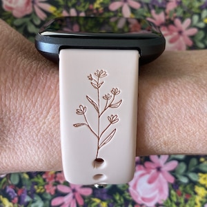 May include: A light pink smartwatch band with a floral design. The band is attached to a black smartwatch. The floral design is a detailed, brown line drawing of a plant with leaves and flowers. The band is secured with a silver clasp.