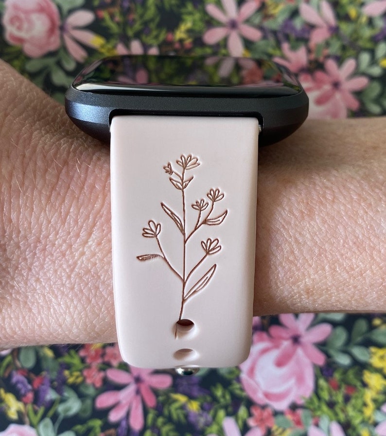 May include: A light pink smartwatch band with a floral design. The band is attached to a black smartwatch. The floral design is a detailed, brown line drawing of a plant with leaves and flowers. The band is secured with a silver clasp.