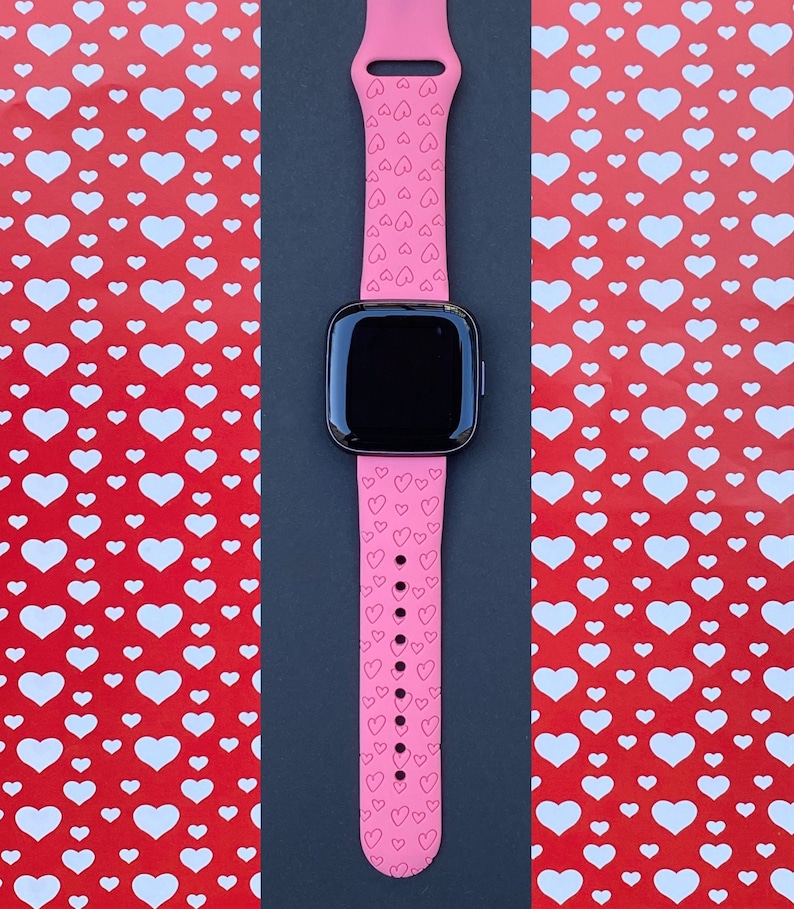 May include: A pink silicone watch strap with a heart pattern. The strap is designed for a smartwatch and has a black screen.