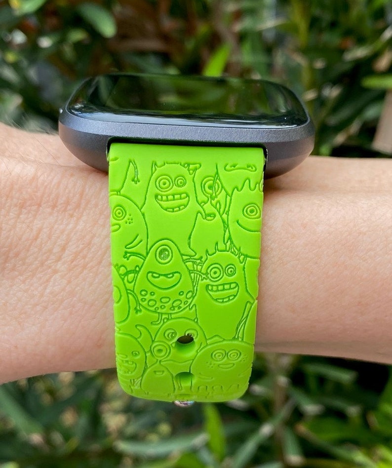 Puede incluir: Una banda de reloj inteligente verde con un dise&ntilde;o de monstruo de dibujos animados. La banda est&aacute; hecha de silicona y tiene una hebilla negra.