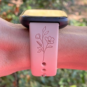 May include: A pink smartwatch band with a floral design. The band has a black watch face and three holes for adjusting the fit.
