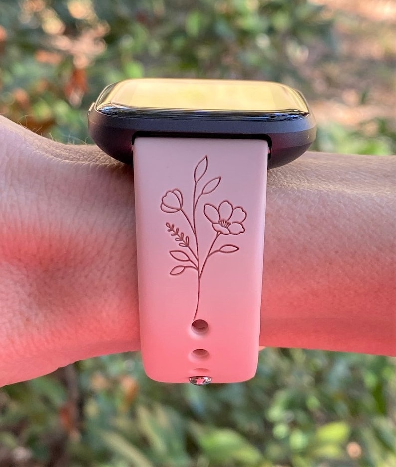 May include: A pink smartwatch band with a floral design. The band has a black watch face and three holes for adjusting the fit.