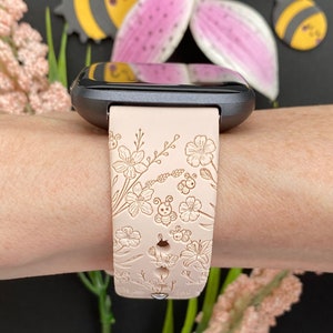 May include: A light pink silicone watch band with a floral and bee design. The band is shown on a person's wrist.