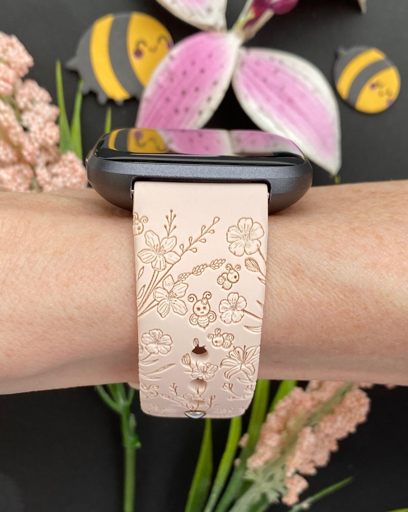 May include: A light pink silicone watch band with a floral and bee design. The band is shown on a person's wrist.