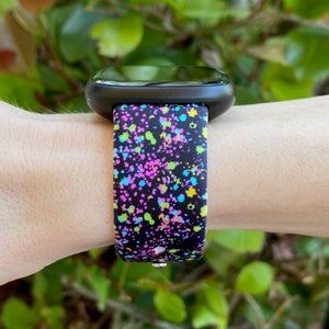 May include: A black smartwatch with a colorful splatter paint design band. The band has a black background with pink, blue, green, and yellow paint splatters.