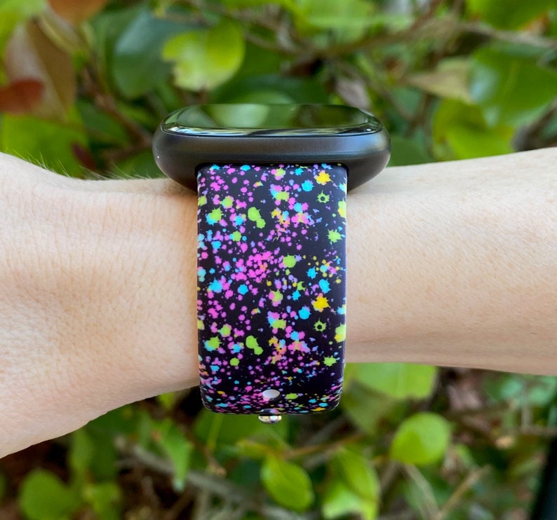 May include: A black smartwatch with a colorful splatter paint design band. The band has a black background with pink, blue, green, and yellow paint splatters.