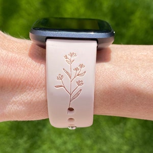 May include: A light pink smartwatch band with a floral design. The band is attached to a dark gray smartwatch. The floral design is a detailed, etched pattern of leaves and flowers.