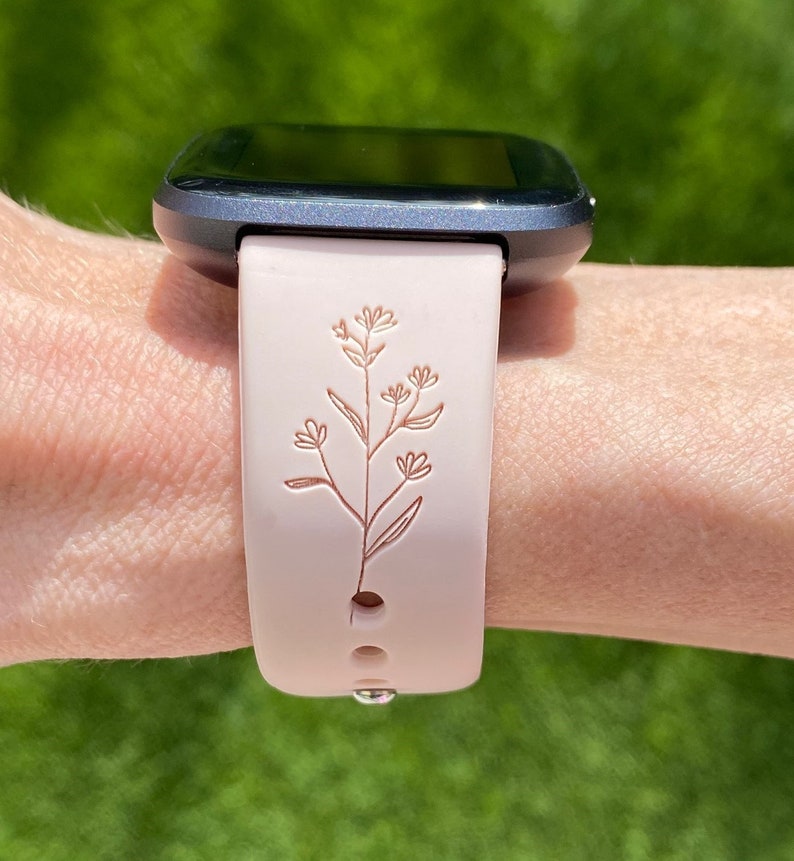 May include: A light pink smartwatch band with a floral design. The band is attached to a dark gray smartwatch. The floral design is a detailed, etched pattern of leaves and flowers.